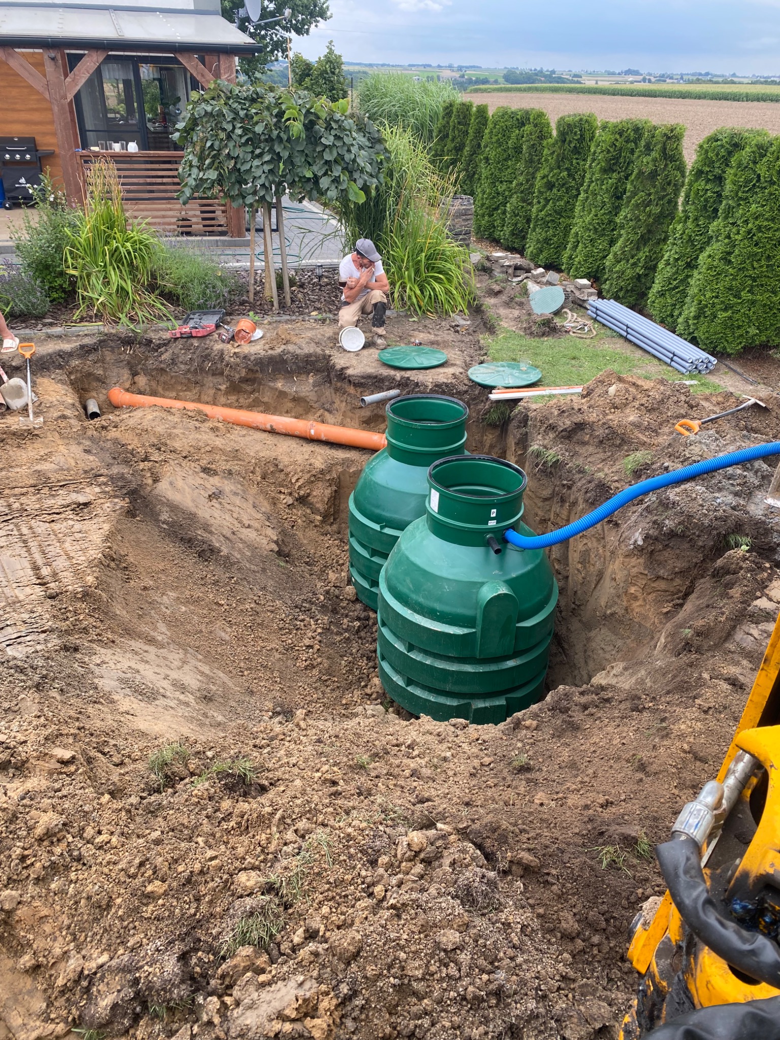 Dwa zielone zbiorniki w wykopie ziemnym, pomarańczowa rura i niebieski wąż, teren budowy z roślinnością w tle. Widoczny pracownik i sprzęt budowlany.
