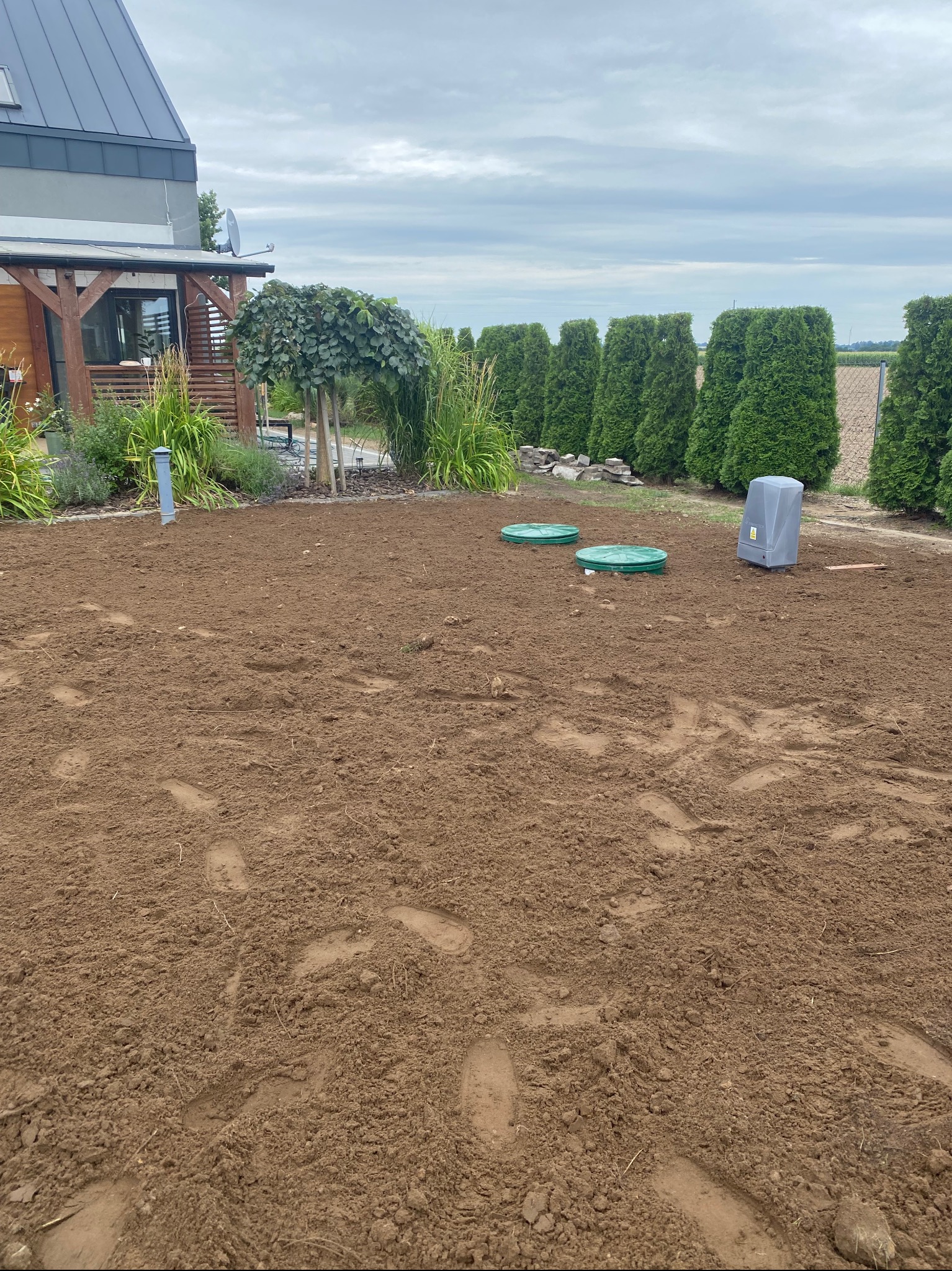 Przygotowany teren pod przydomową oczyszczalnię ścieków z widocznymi studzienkami i zbiornikiem w szarej obudowie. W tle rząd tuj i dom.