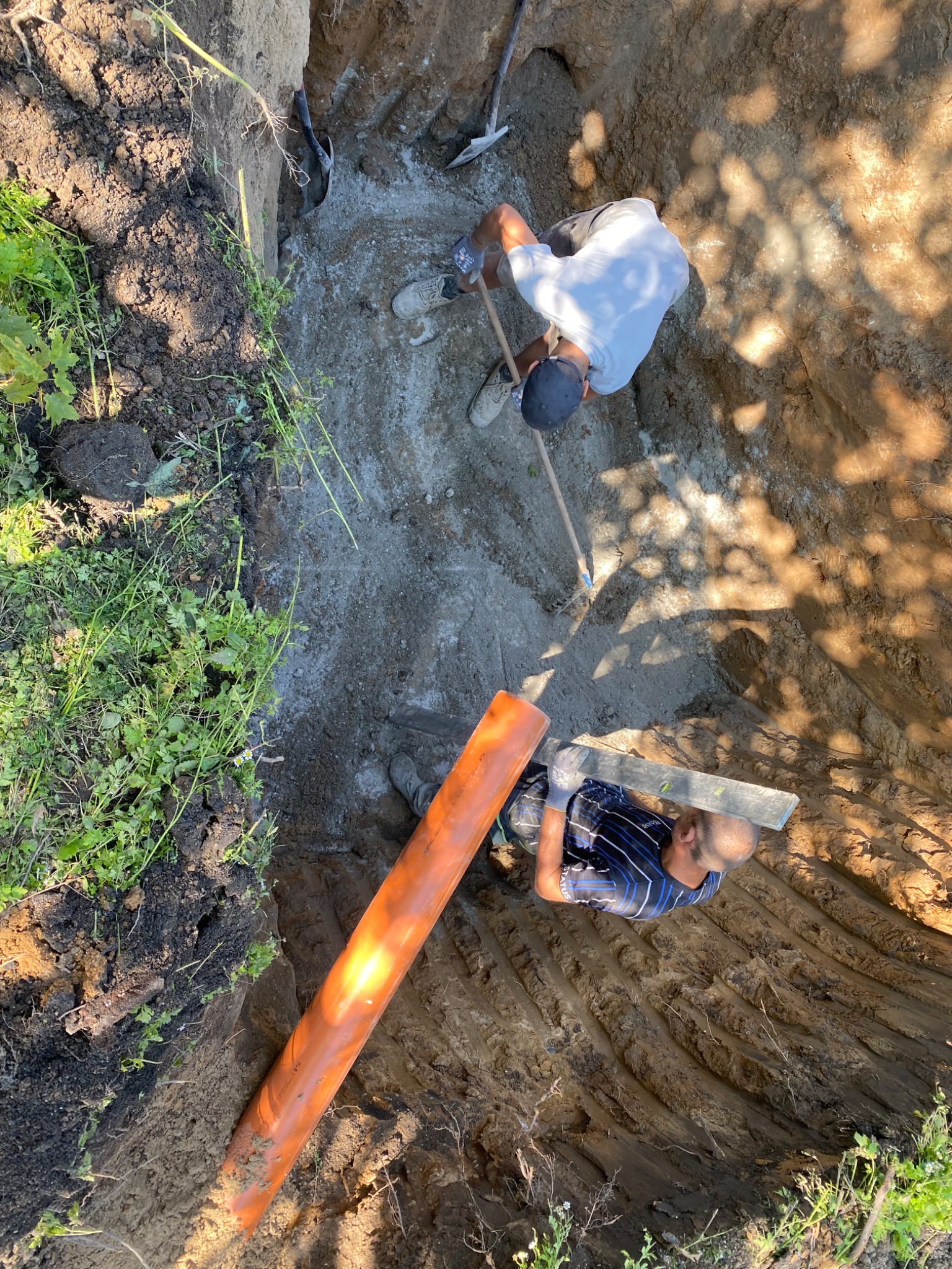 Dwaj mężczyźni w wykopie układają pomarańczową rurę kanalizacyjną, używając poziomicy. Widok z góry, narzędzia i ziemia w tle.