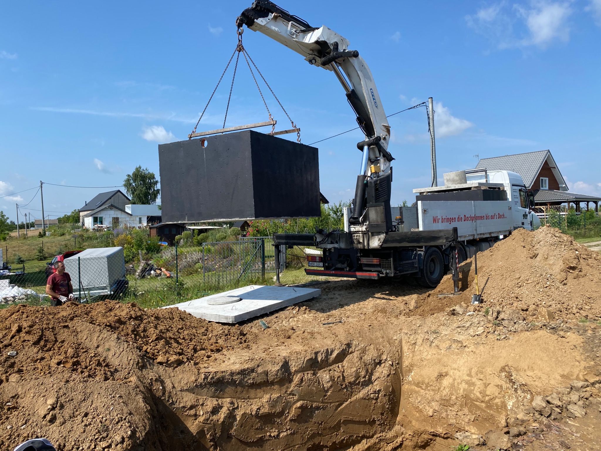 Czarny, betonowy zbiornik zawieszony nad wykopem, transportowany dźwigiem na tle krajobrazu wiejskiego z domami i zielenią. Widoczny pracownik i fragment ciężarówki.