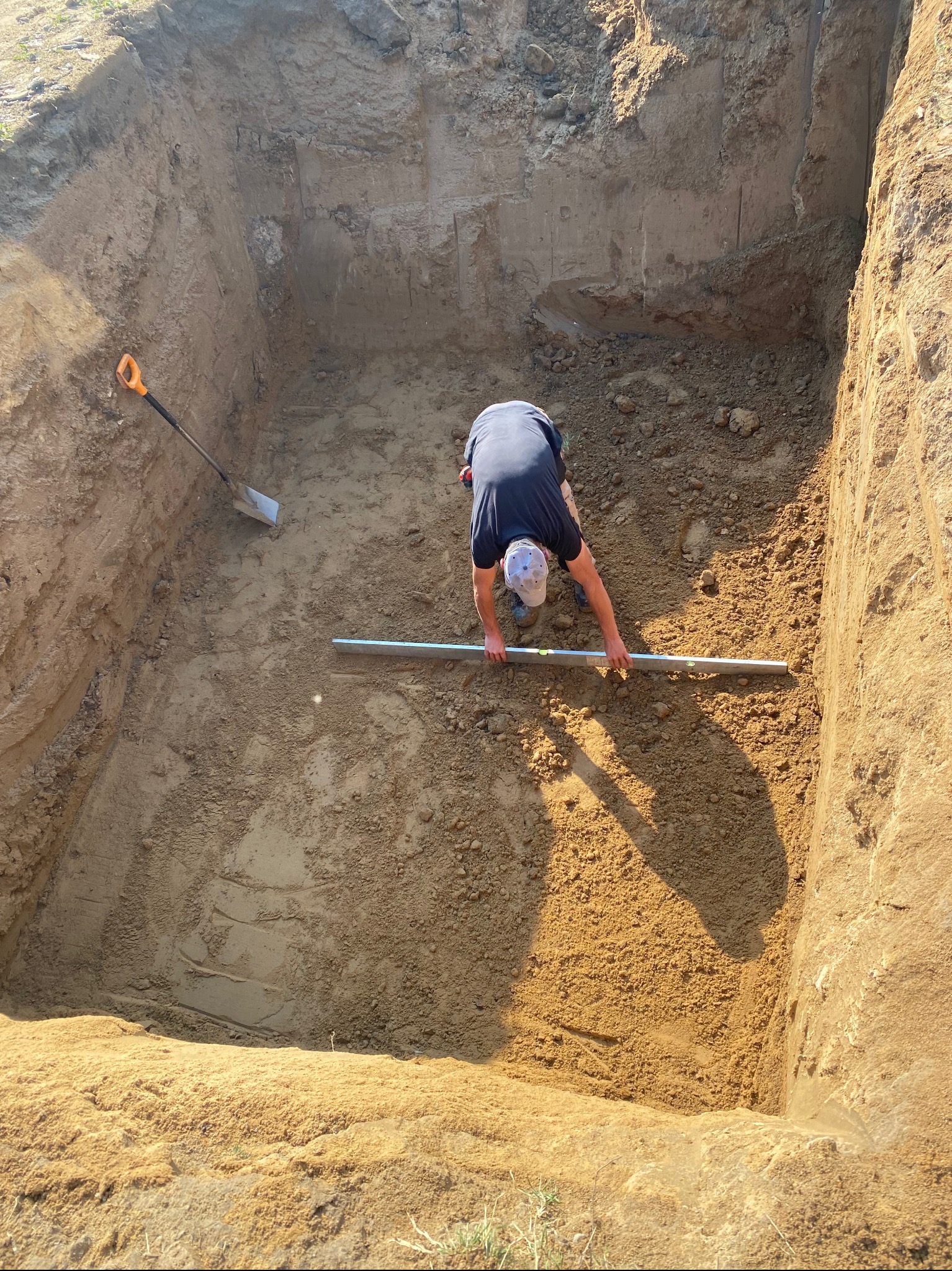 Wykop pod szambo betonowe: mężczyzna poziomuje dno wykopu za pomocą poziomicy, łopata oparta o ścianę wykopu. Słoneczny dzień.
