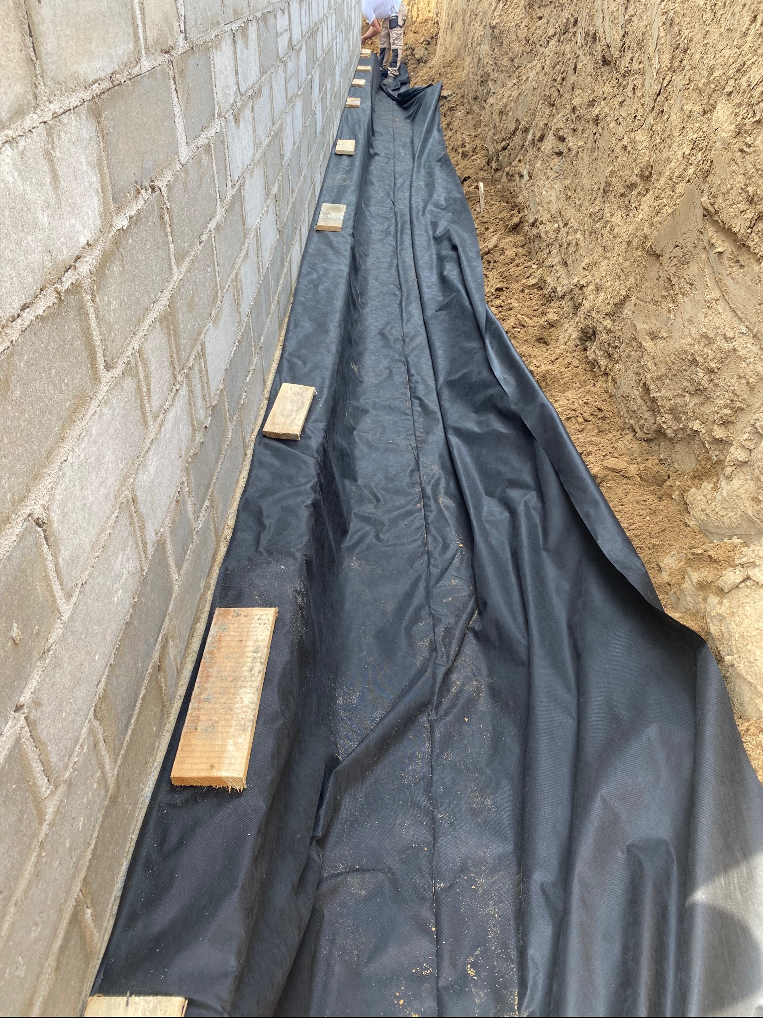 Wykop z izolacją pionową przy fundamencie budynku z bloczków betonowych. Czarna folia zabezpieczająca, drewniane rozpórki. Prace ziemne w toku, widoczny fragment osoby.