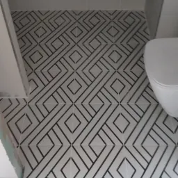 Wzorzysta, biało-czarna podłoga w łazience z płytkami ułożonymi w geometryczny deseń, widoczna częściowo toaleta i szafka pod umywalką, z fragmentem podłogi drewnopodobnej przed wejściem.