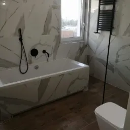 Biała wanna obudowana płytkami imitującymi marmur, czarna armatura łazienkowa, drewnopodobna podłoga z płytek, grzejnik drabinkowy w kolorze czarnym i biała toaleta w nowoczesnej łazience.