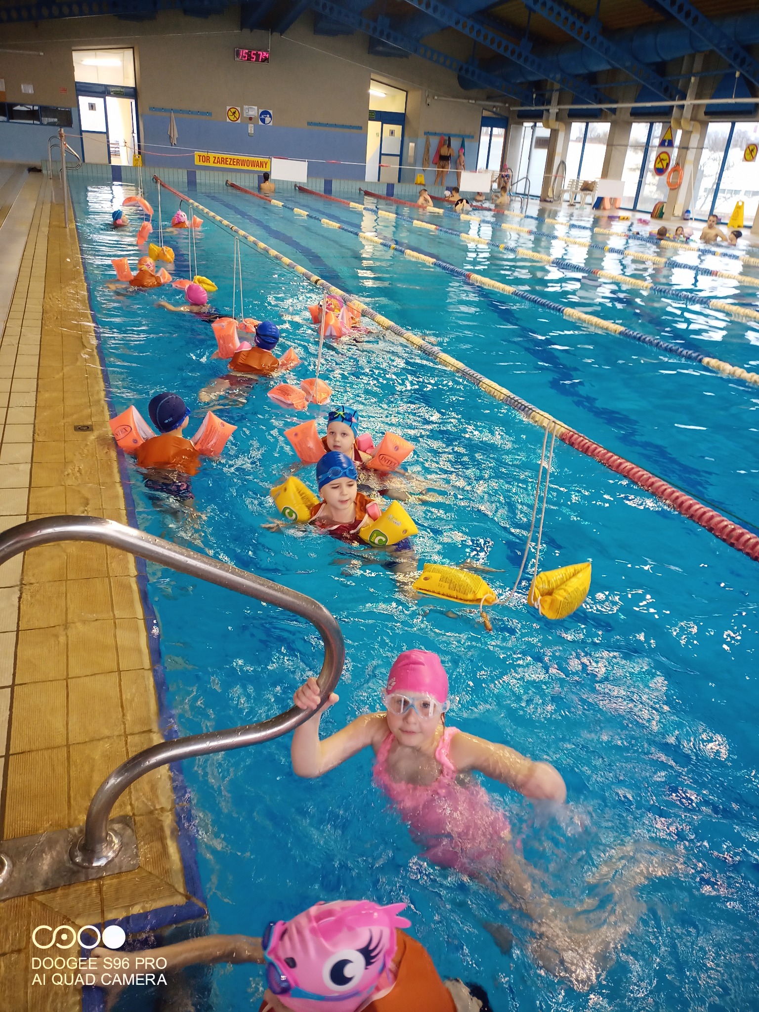 Grupa dzieci w różowych i pomarańczowych rękawkach pływa w basenie krytym, jedna dziewczynka w różowym czepku trzyma się metalowej poręczy na pierwszym planie.