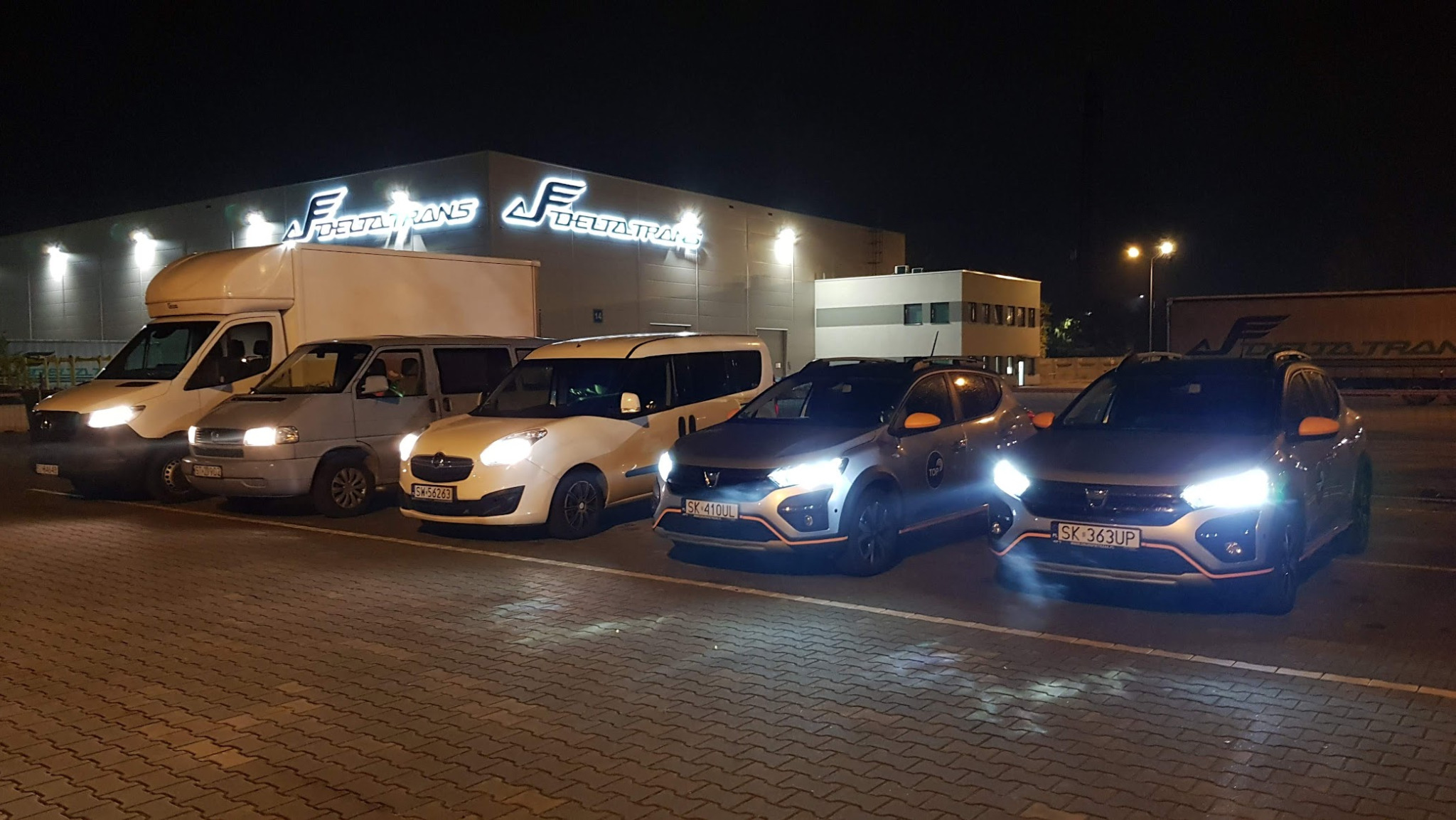 Flota pojazdów dostawczych i osobowych na parkingu firmy Delta Trans nocą, z włączonymi światłami przed budynkiem z logo firmy.