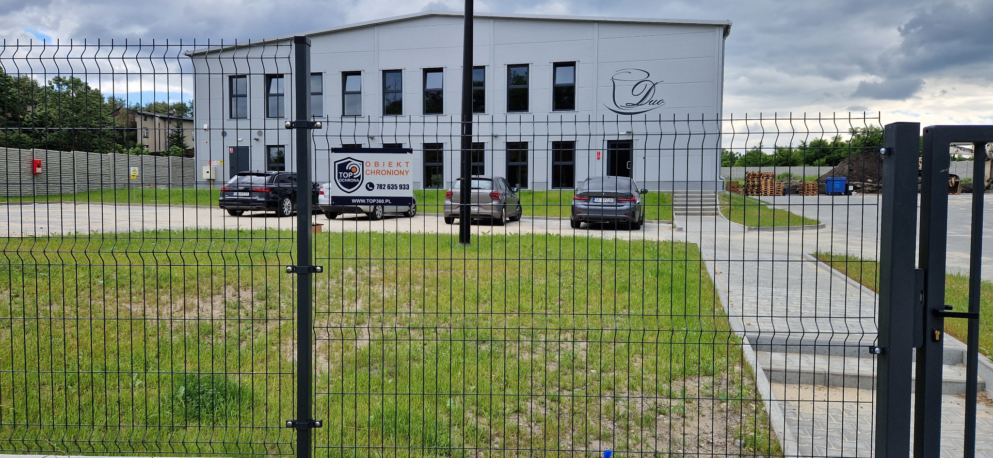 Ogrodzony teren z budynkiem firmy z logo, zaparkowanymi samochodami i tablicą informacyjną 'Obiekt Chroniony'.