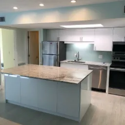 Nowoczesna, jasna kuchnia z wyspą i blatem z granitu, białymi szafkami, stalową lodówką i sprzętem AGD, wykończona w odcieniach bieli i błękitu.