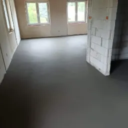 Świeżo wylana, gładka posadzka w surowym wnętrzu z oknami i ścianą z betonu komórkowego.