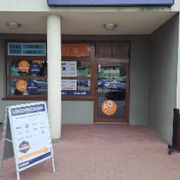 Fasada placówki Unilink Ubezpieczenia z witrynami oklejonymi informacjami o ubezpieczeniach i kredytach, na chodniku przed wejściem ustawiony stojak reklamowy z ofertą ubezpieczeń.