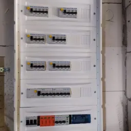 Rozdzielnica elektryczna z zamontowanymi bezpiecznikami i zabezpieczeniami przepięciowymi, widoczna na tle surowej ściany z bloczków.