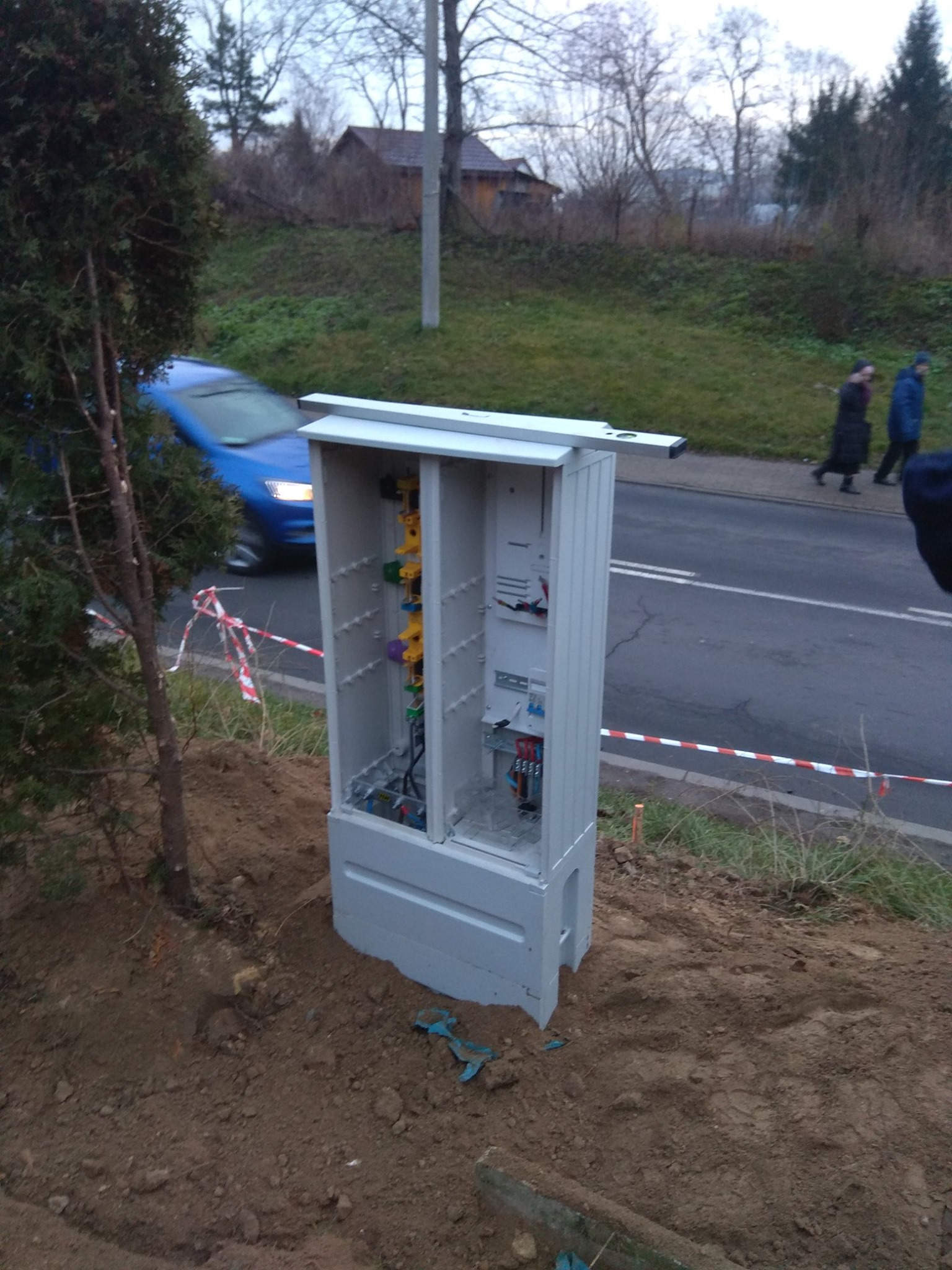Otwarta, szara skrzynka elektryczna z widocznymi przewodami i elementami instalacji, umieszczona na ziemi obok ulicy z przejeżdżającym niebieskim samochodem i pieszymi w tle.
