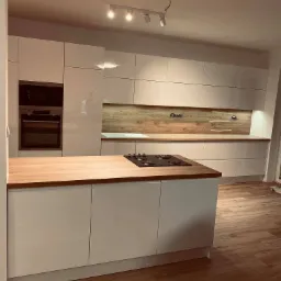 Nowoczesna kuchnia z białymi, lakierowanymi frontami, drewnianym blatem i panelem ściennym, oświetleniem punktowym i wbudowanym piekarnikiem.