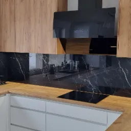 Nowoczesna kuchnia z drewnianymi szafkami, czarnym panelem ściennym z efektem marmuru, blatem imitującym drewno i widokiem na zaśnieżony krajobraz za oknem.