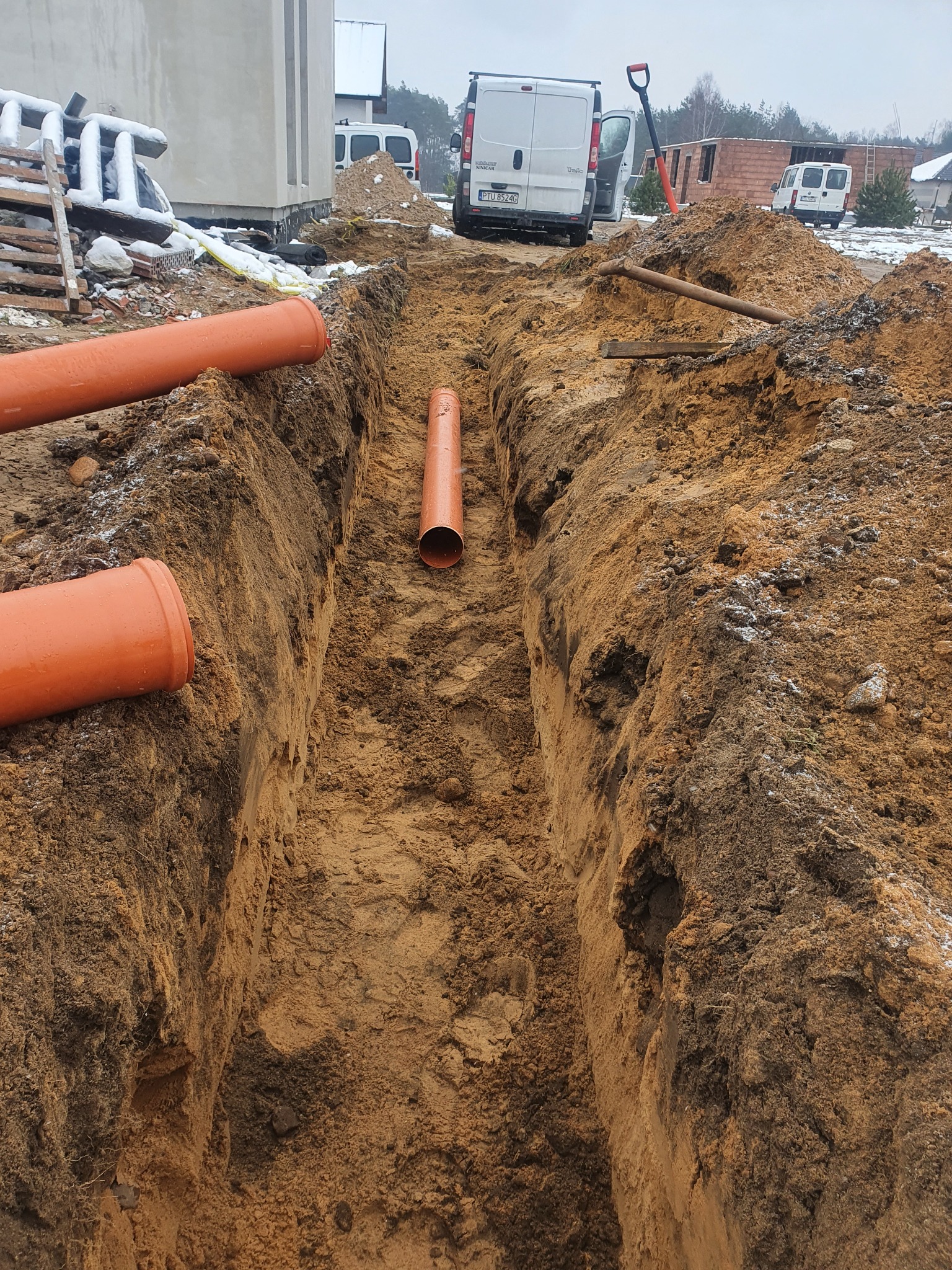 Wykop z pomarańczowymi rurami kanalizacyjnymi w ziemi, widoczne samochody dostawcze w tle, teren budowy.