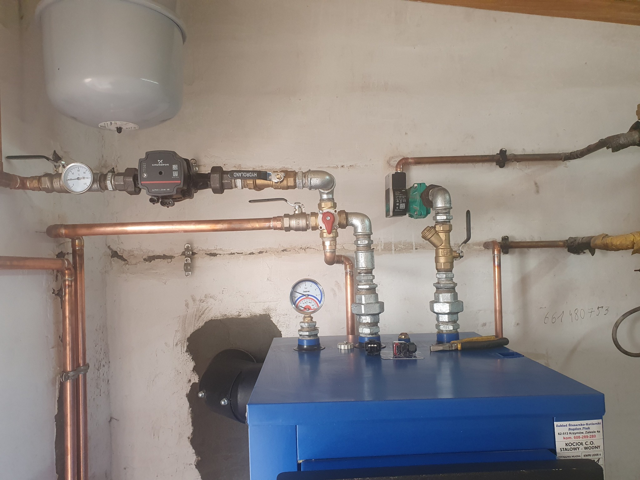 Niebieski kocioł C.O. stalowy-wodny z podłączonymi miedzianymi rurami, manometrem i pompą obiegową Grundfos Alpha1, zamontowany na tle ściany z widocznymi śladami napraw i instalacji.