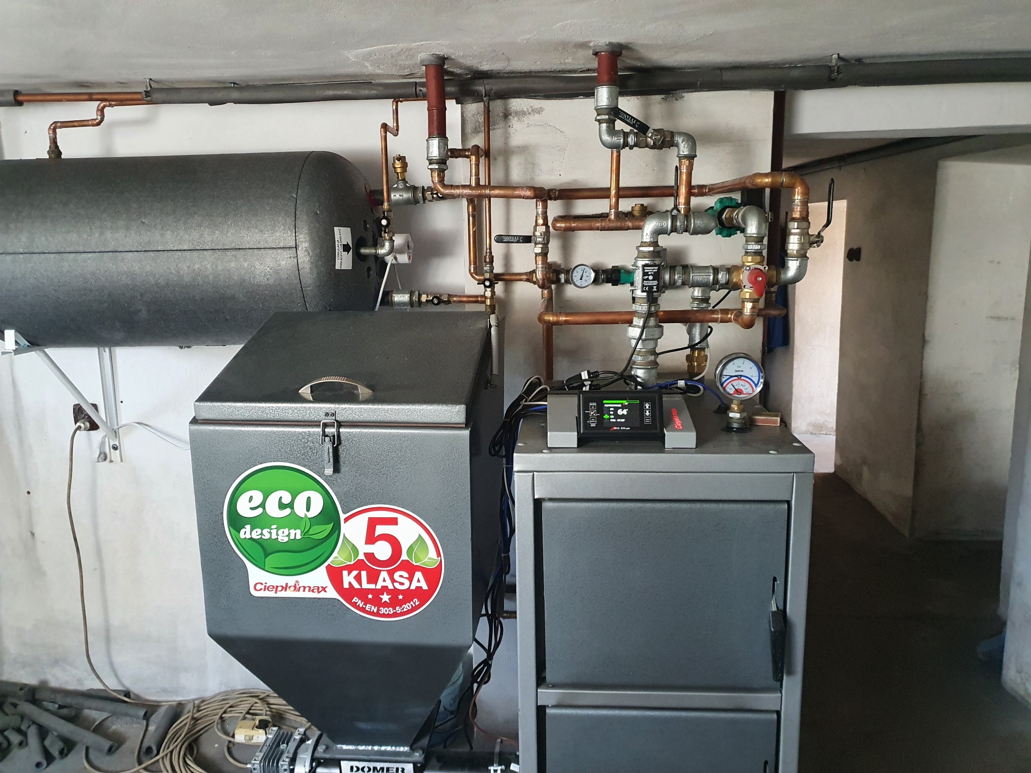 Kotłownia z piecem na paliwo stałe z podajnikiem, zbiornikiem buforowym i instalacją hydrauliczną z miedzianymi rurami oraz automatyką sterującą, widoczne oznaczenia klasy energetycznej i eco design.