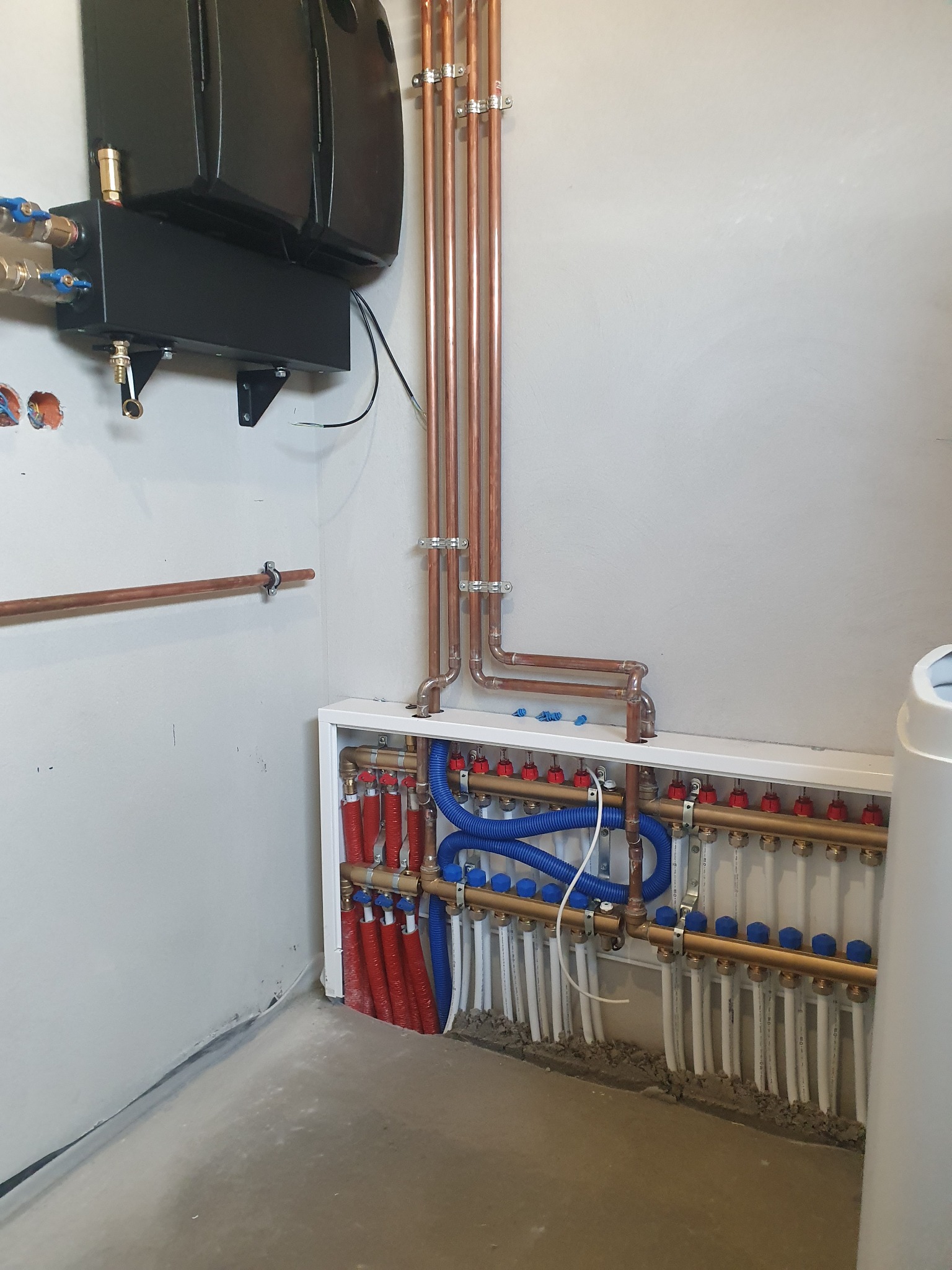 Rozdzielacz ogrzewania podłogowego z zaworami, rurami miedzianymi i niebieskimi przewodami, zamontowany w pomieszczeniu z surową wylewką betonową na podłodze.