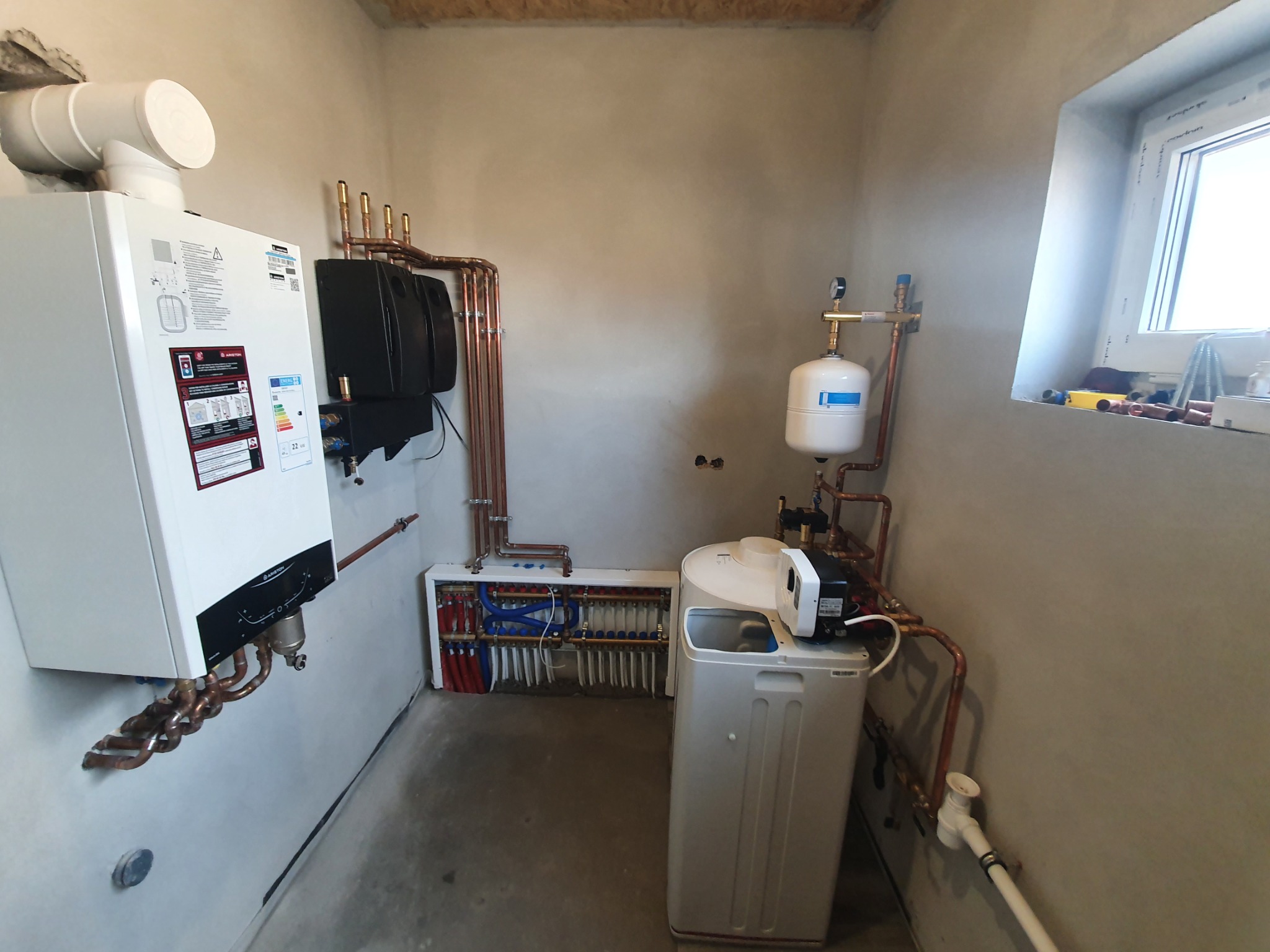 Nowoczesna kotłownia z białym piecem gazowym, rozdzielaczem podłogowym, zasobnikiem CWU i zmiękczaczem wody, połączone miedzianą instalacją w surowym pomieszczeniu z oknem.