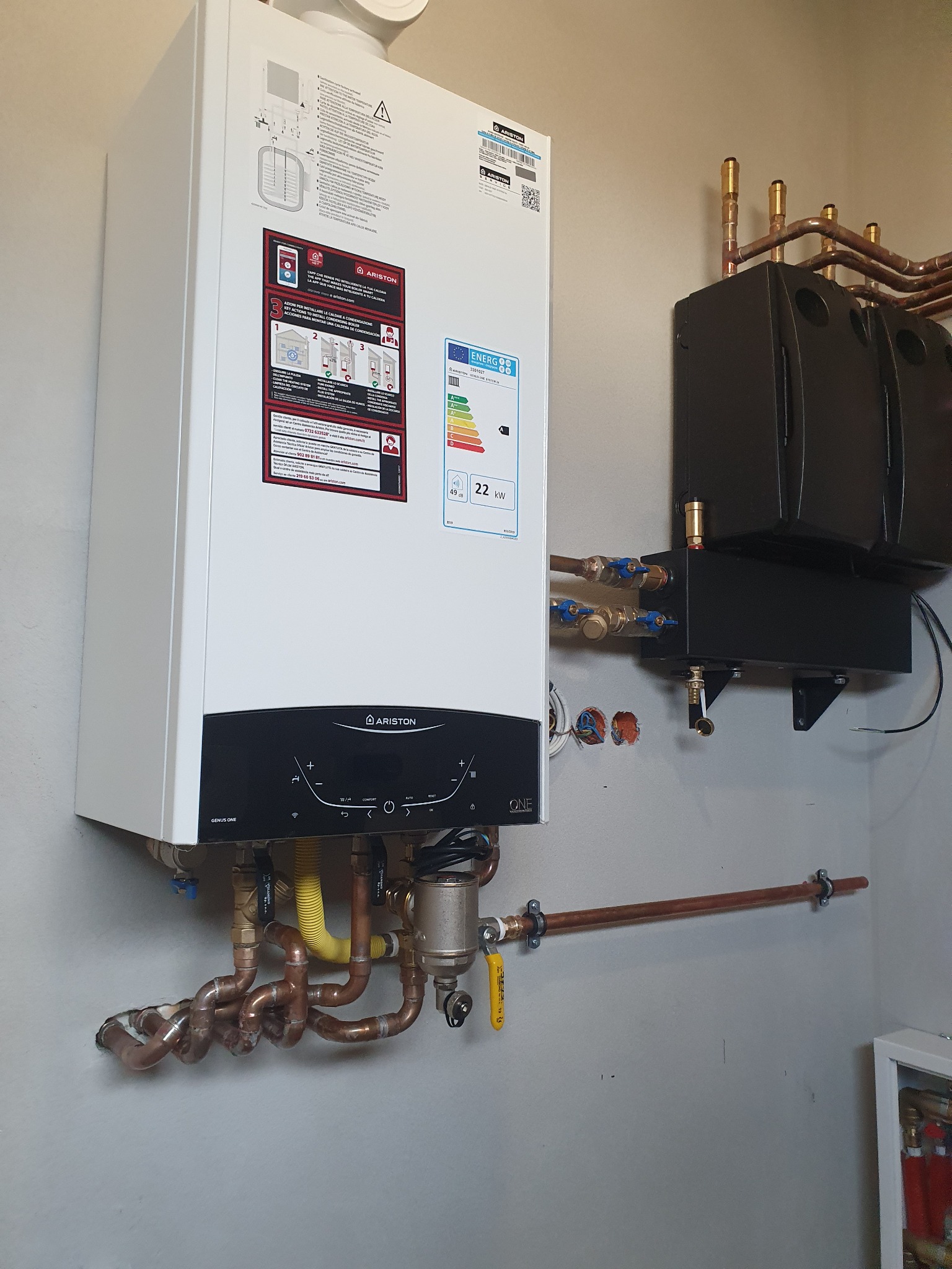 Biały piec gazowy marki Ariston zamontowany na szarej ścianie, z widocznymi miedzianymi rurami, filtrem i elementami instalacji, obok zamontowany czarny zbiornik wyrównawczy.