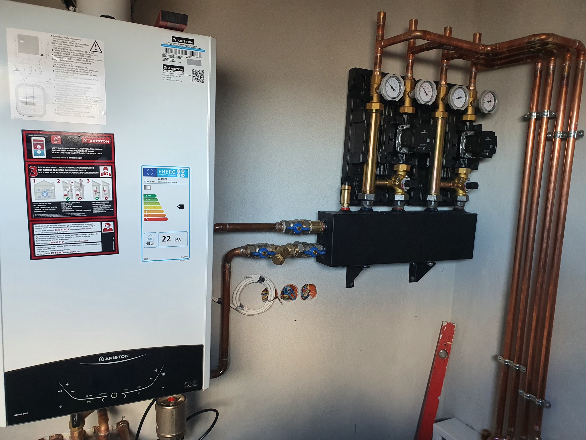 Biały piec gazowy marki Ariston z czarnym panelem sterowania, zamontowany na ścianie obok rozdzielacza z czterema pompami obiegowymi i manometrami, połączonego miedzianymi rurami, widoczne...