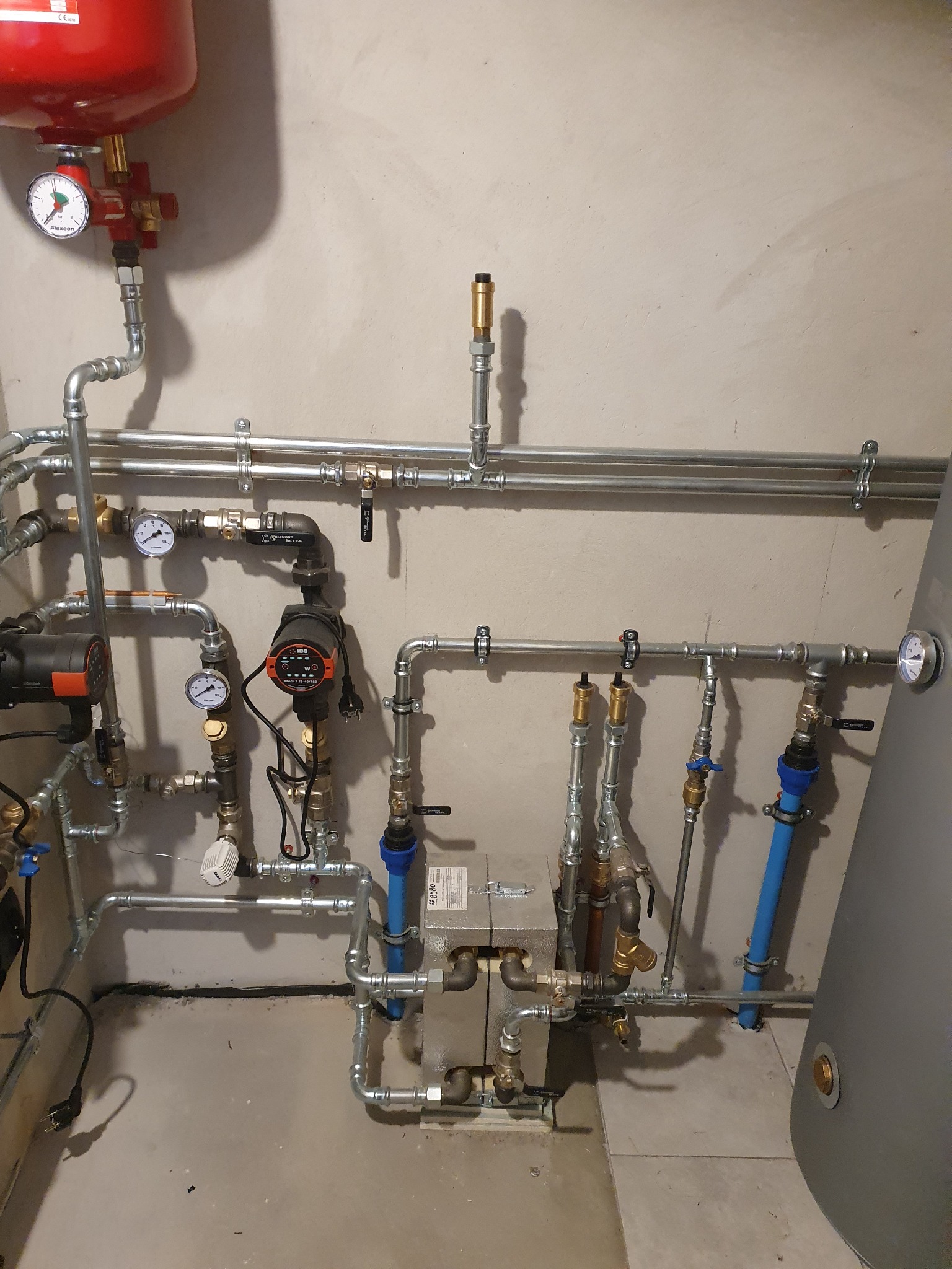 Rozbudowana instalacja grzewcza w piwnicy z licznymi rurami, zaworami, pompami obiegowymi i zbiornikiem wyrównawczym, z widocznymi manometrami i etykietami urządzeń.