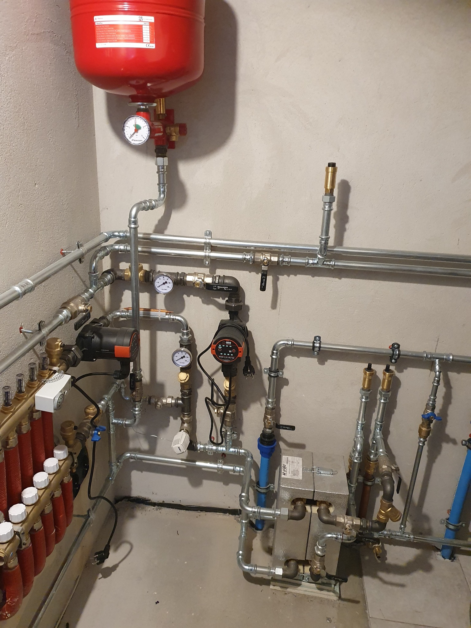 Kompleksowa instalacja grzewcza z licznymi rurami, zaworami, pompami obiegowymi, manometrami i czerwonym zbiornikiem wyrównawczym, zamontowana przy szarej ścianie.