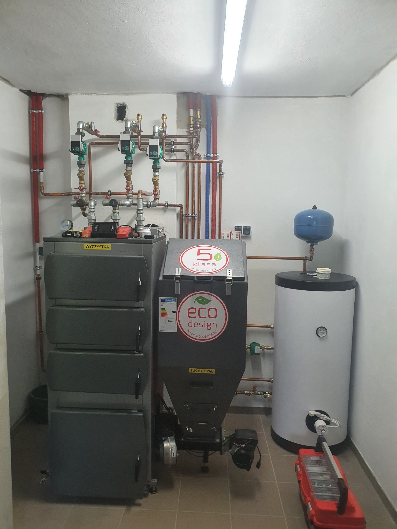 Kotłownia z dwoma piecami, jeden z podajnikiem, zasobnik ciepłej wody i rozbudowana instalacja z rur miedzianych, widoczna skrzynka narzędziowa na posadzce.