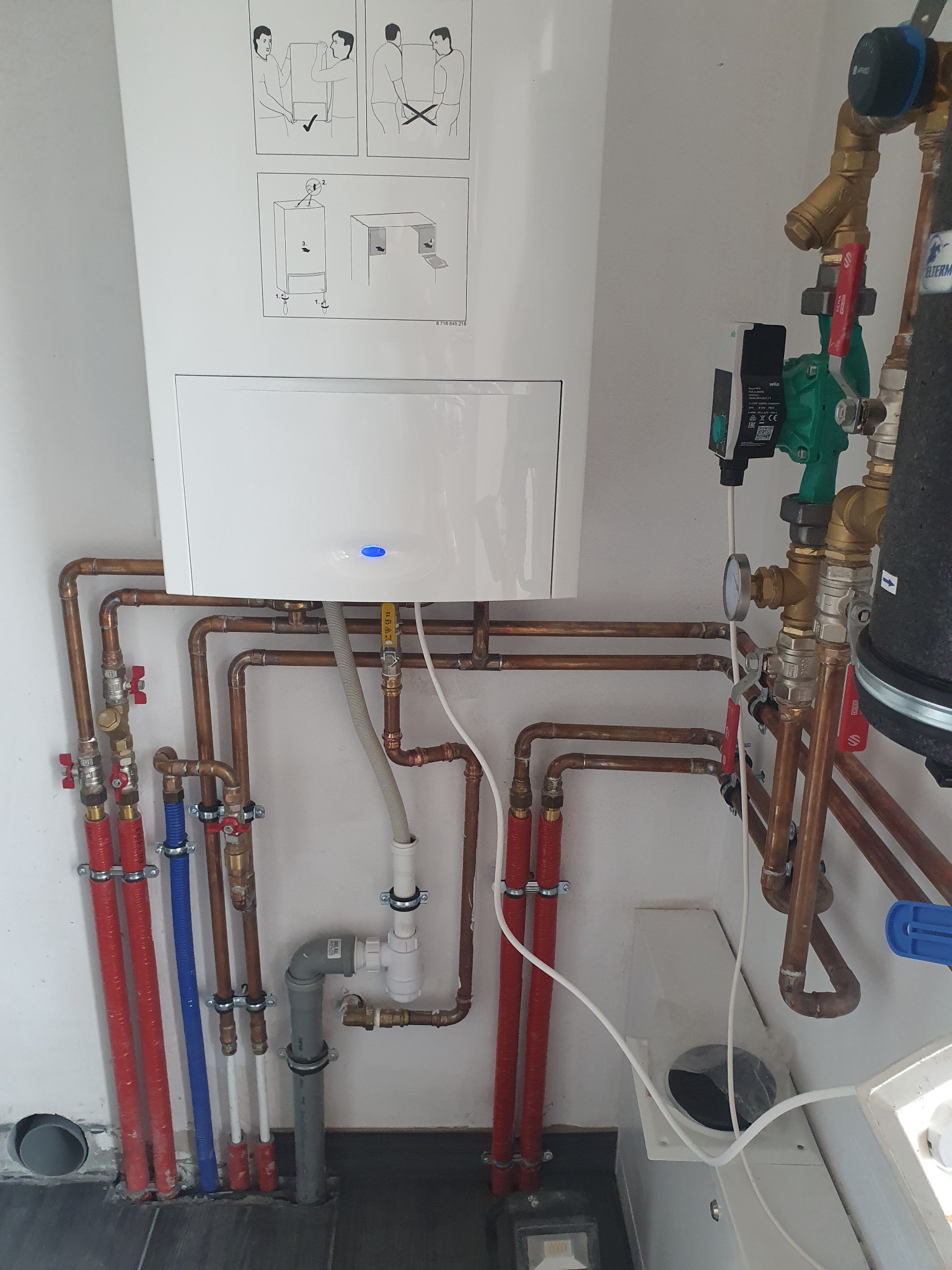 Biały piec CO z niebieską diodą, zamontowany na ścianie z instalacją z miedzianych rur, zaworami z czerwonymi uchwytami, pompą obiegową Wilo i schematem instruktażowym na obudowie.