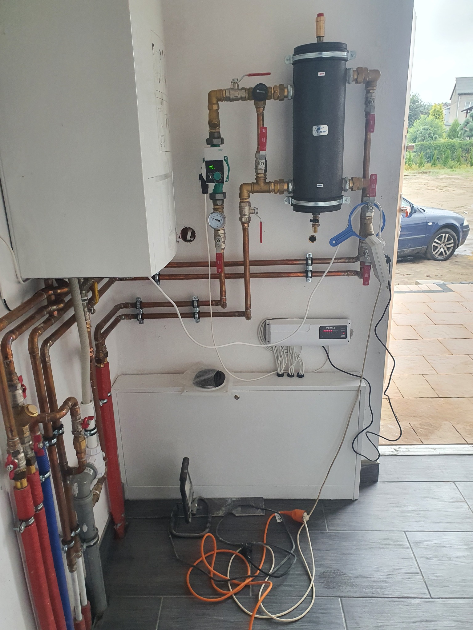 Zainstalowana kotłownia z piecem, zasobnikiem ciepłej wody i rozbudowaną instalacją hydrauliczną z licznymi rurami miedzianymi i zaworami, widoczny sterownik, na podłodze leżą kable i lampa robocza.