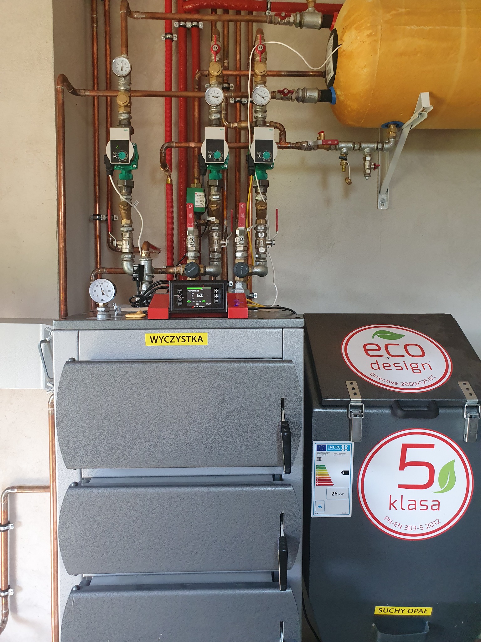 Instalacja grzewcza z kotłem na paliwo stałe, widoczne miedziane rury, zawory, pompy obiegowe, manometry, sterownik elektroniczny z wyświetlaczem, zbiornik wyrównawczy, etykieta 'Eco Design'...