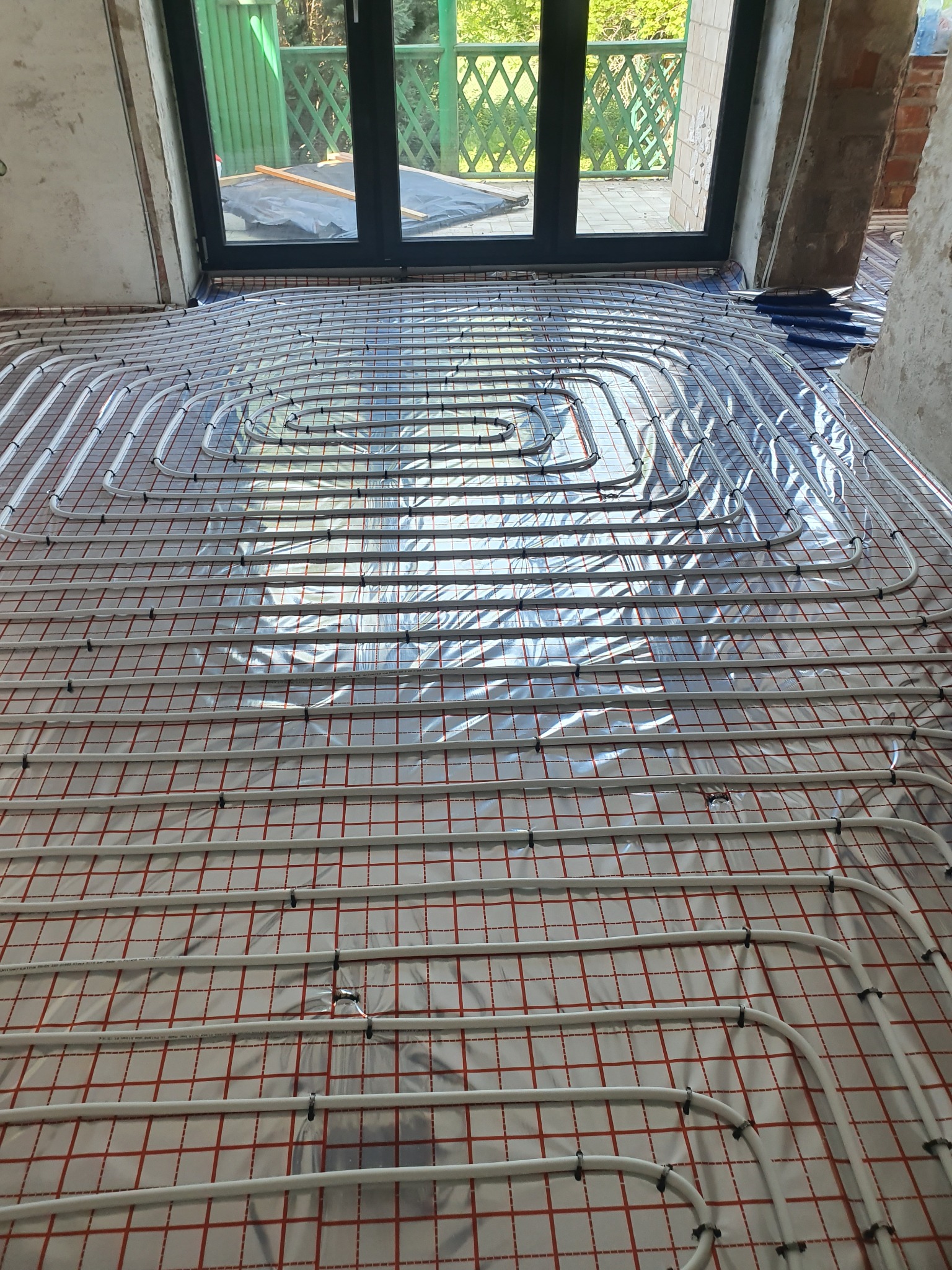 Białe rury ogrzewania podłogowego ułożone spiralnie na folii z czerwoną siatką, widoczne czarne klipsy mocujące, w tle okno balkonowe i surowe ściany w trakcie remontu.