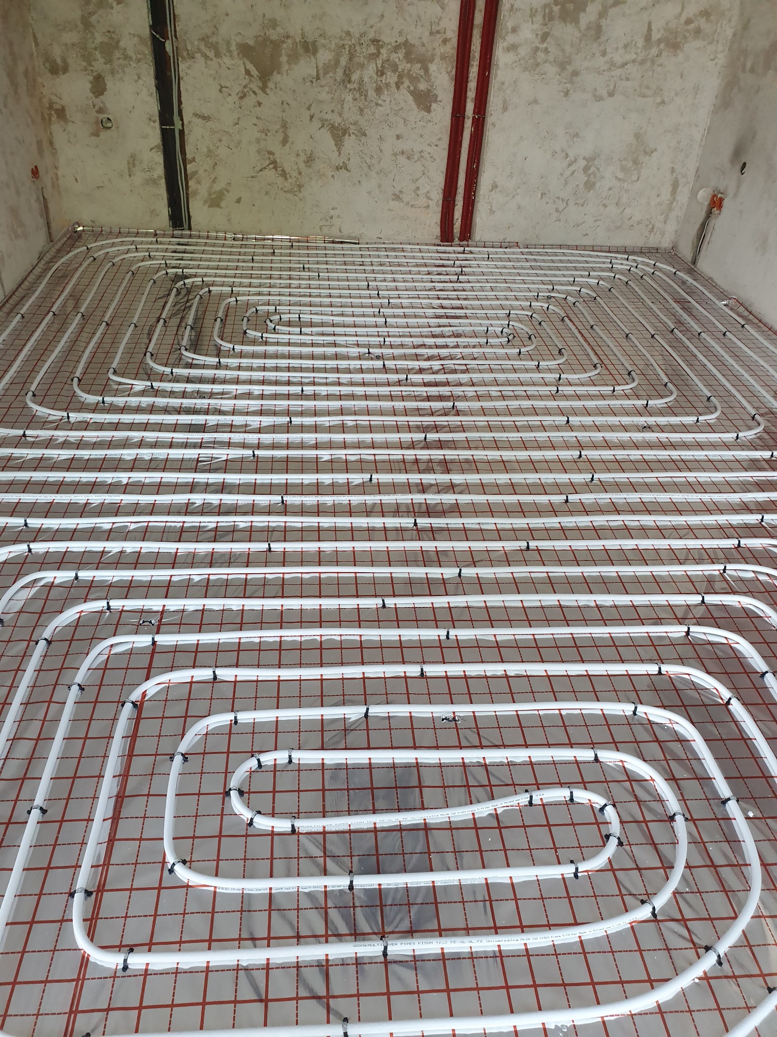 Rozłożone białe rury ogrzewania podłogowego, przymocowane czarnymi uchwytami do czerwonej siatki na podłodze w surowym stanie.
