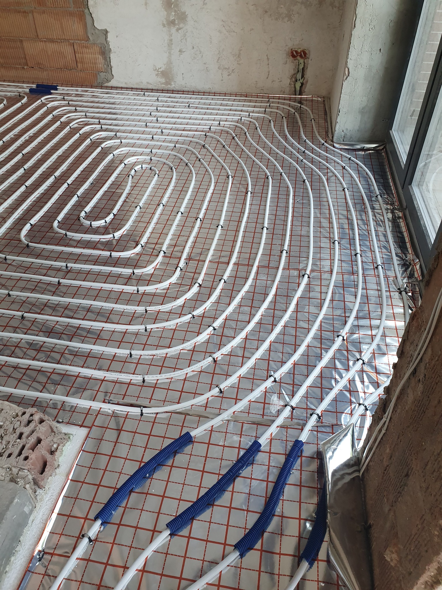 Montaż ogrzewania podłogowego: białe rury ułożone spiralnie na folii izolacyjnej z czerwoną siatką, zabezpieczone czarnymi zaciskami, w surowym wnętrzu z widoczną cegłą i fragmentem okna.