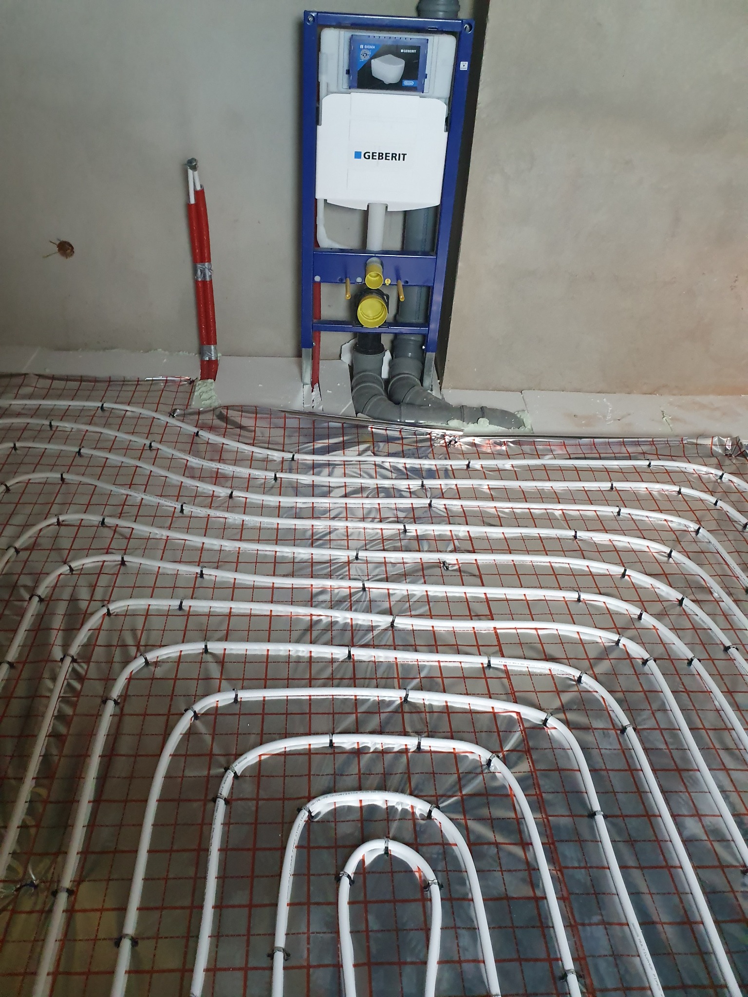 Białe rury ogrzewania podłogowego, przymocowane czarnymi klipsami do czerwonej siatki na folii, instalacja podtynkowa Geberit z szarą rurą kanalizacyjną w tle, czerwona rura pionowa przy ścianie.