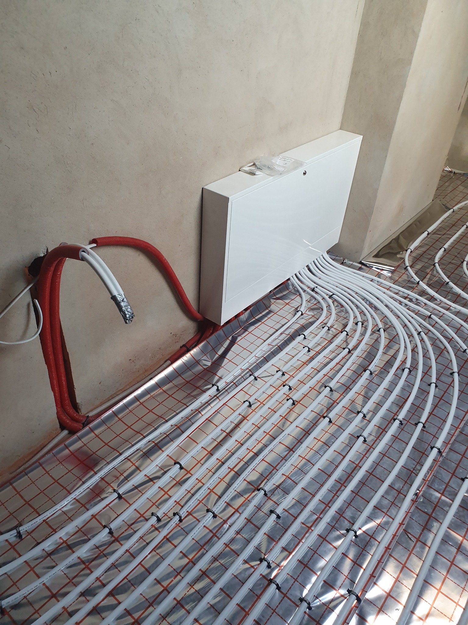 Instalacja ogrzewania podłogowego z białymi rurkami na folii refleksyjnej z czerwoną siatką, widoczne czerwone rury w peszlu i biała szafka rozdzielcza na ścianie w surowym stanie.