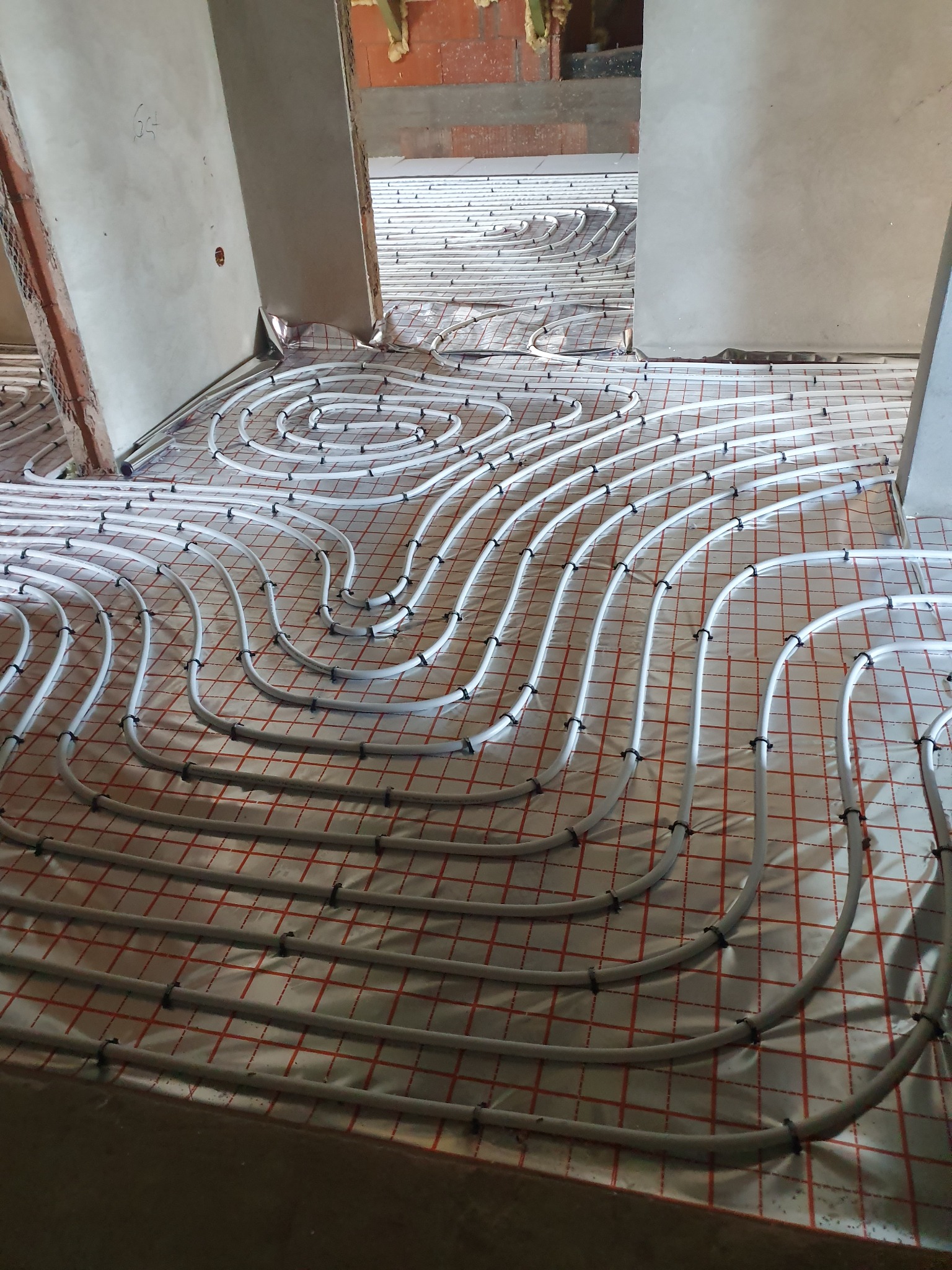 Rozłożone, białe rury ogrzewania podłogowego, przymocowane czarnymi klipsami do folii z czerwoną kratką, widok na surowy stan pomieszczenia w trakcie remontu.
