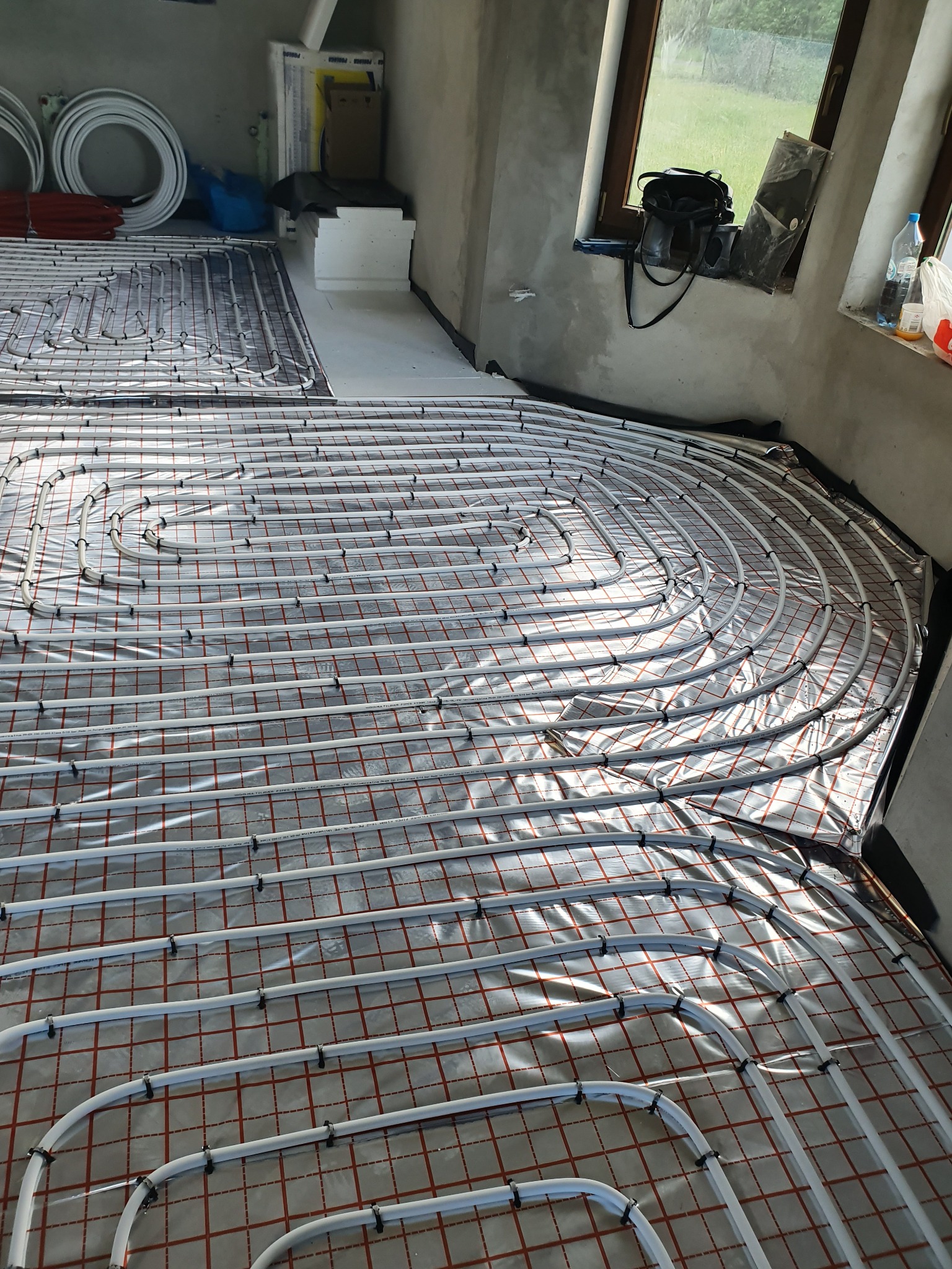 Rozłożona instalacja ogrzewania podłogowego z białych rur na srebrnej folii z czerwoną kratką, mocowana czarnymi klipsami w surowym wnętrzu budynku z oknem.