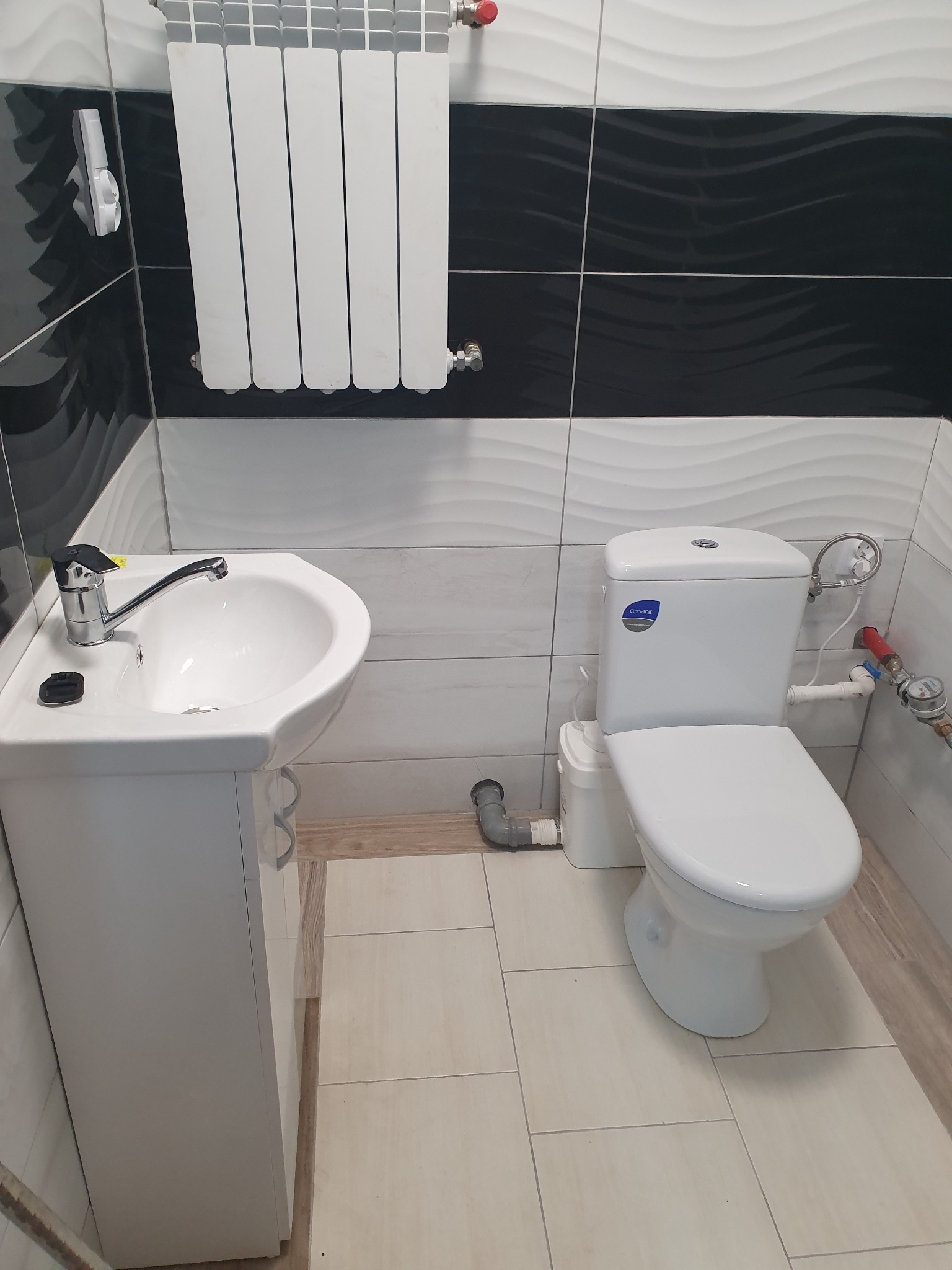 Wykończona łazienka z białą umywalką na szafce, toaletą z kompaktowym zbiornikiem, grzejnikiem panelowym i czarno-białymi płytkami ściennymi z motywem fal. Widoczny wodomierz i instalacja...