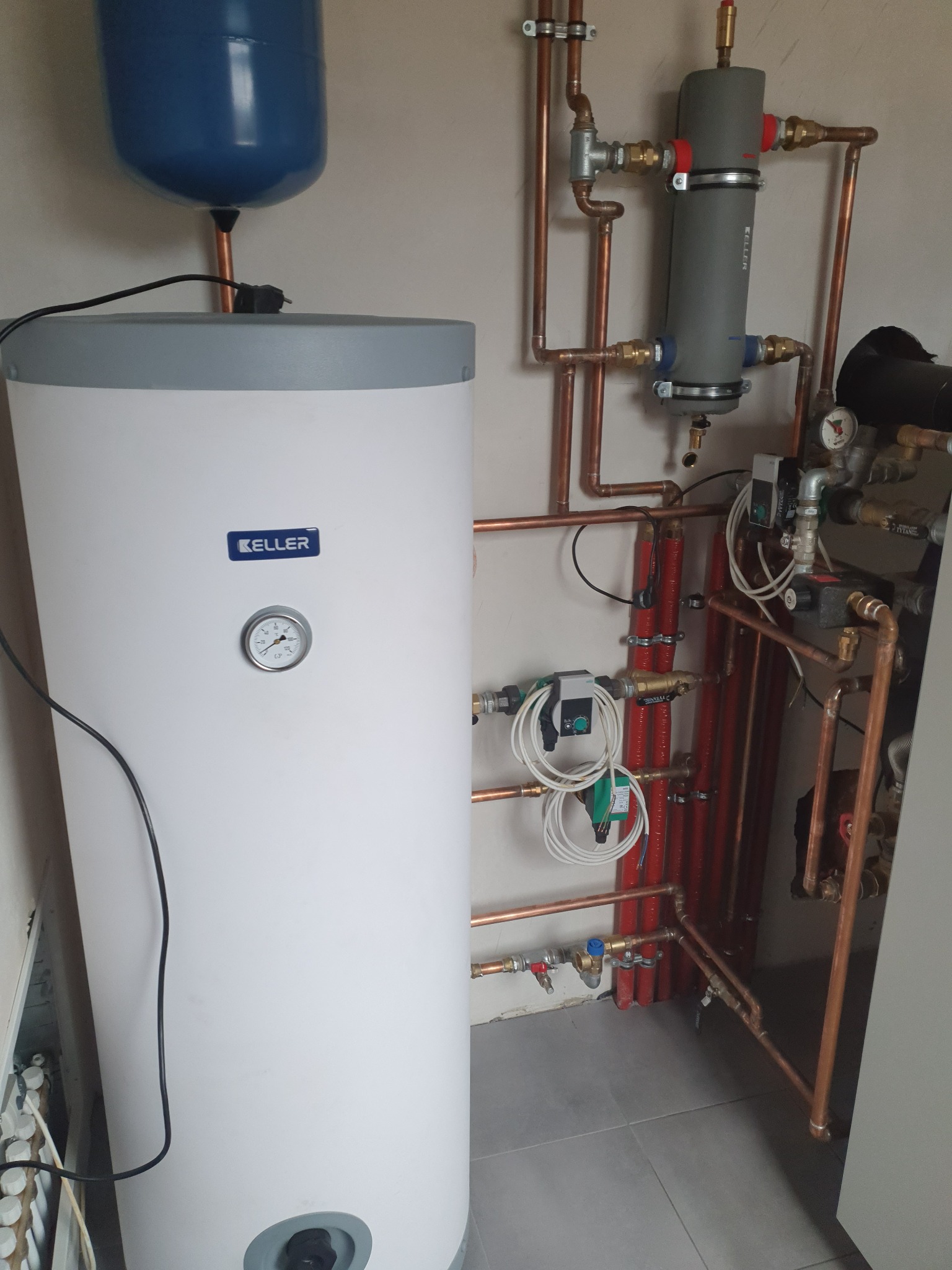 Biały zasobnik ciepłej wody marki Keller z niebieskim zbiornikiem wyrównawczym, połączony miedzianymi rurami i zaworami w kotłowni, widoczne elementy automatyki i manometr.