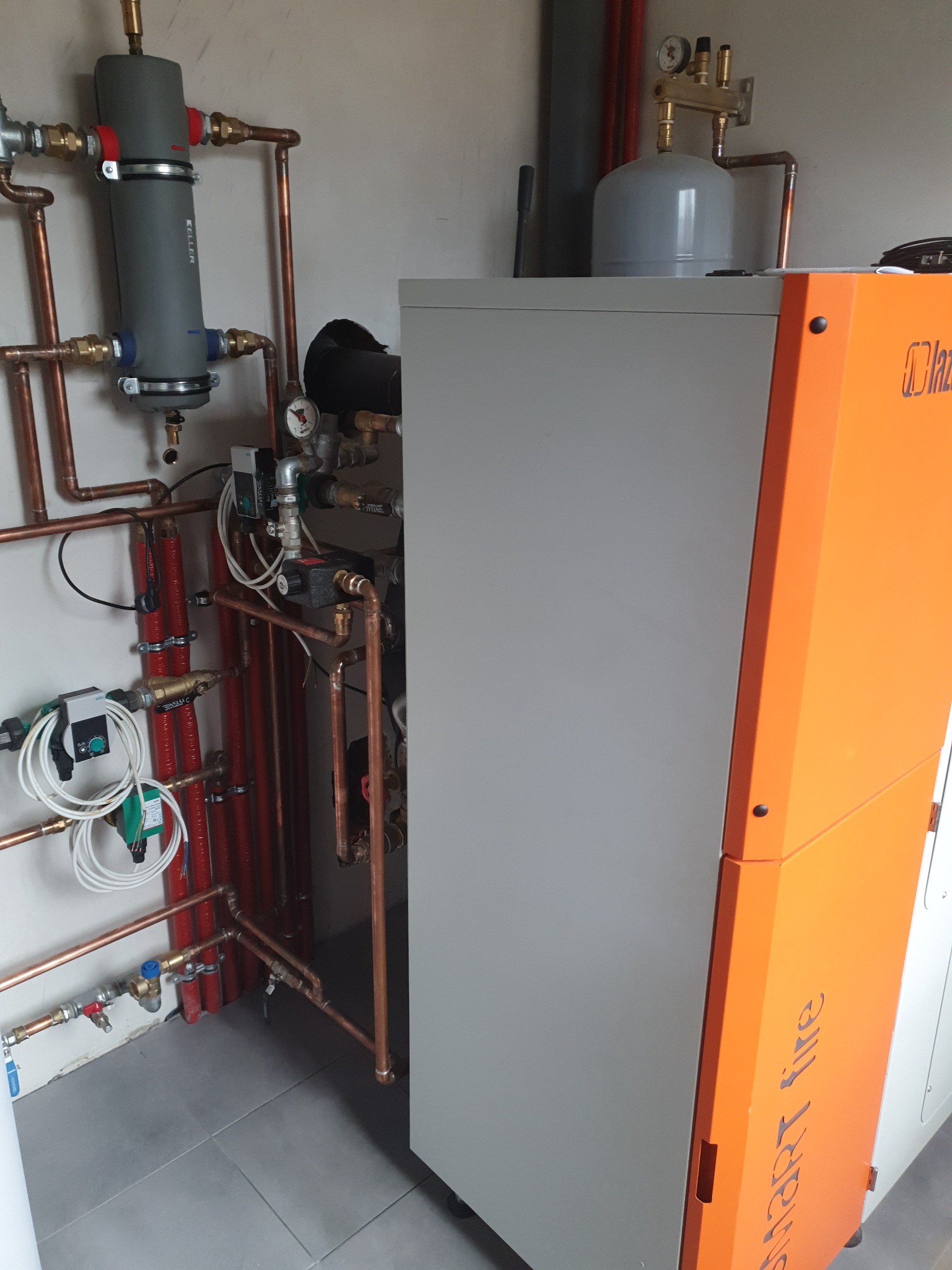 Pomarańczowo-szary piec grzewczy z podłączoną instalacją hydrauliczną z miedzianych rur, widoczne liczniki i zawory.