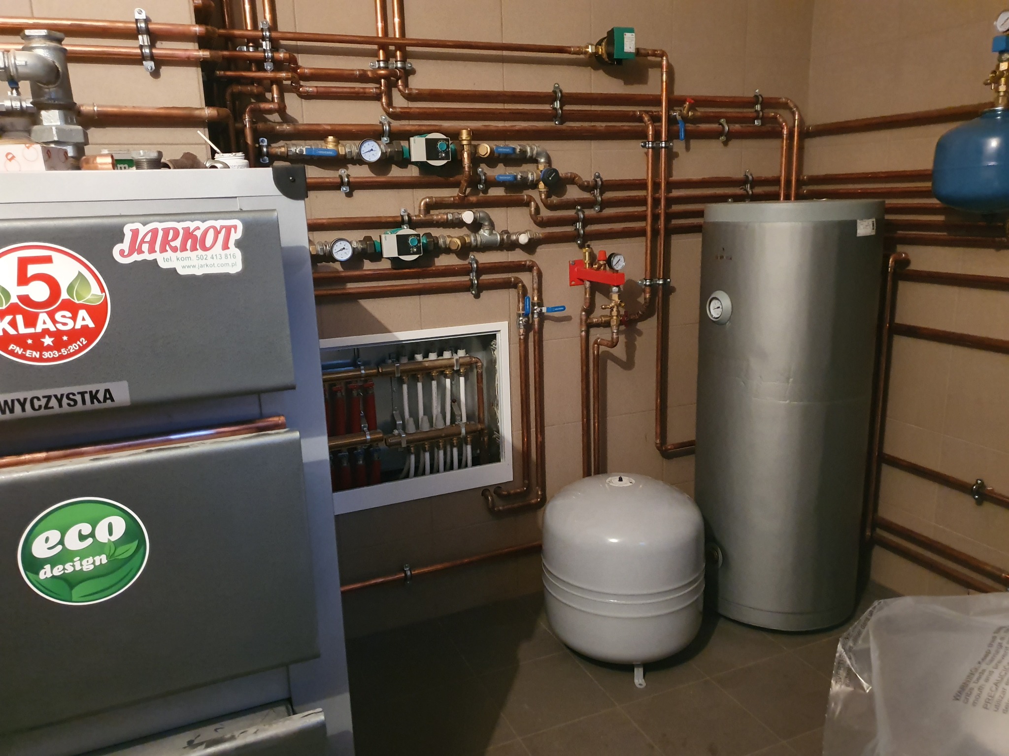 Kotłownia z kotłem na paliwo stałe marki Jarkot, zbiornikami akumulacyjnymi i rozbudowaną instalacją hydrauliczną z miedzianych rur, widoczne naklejki informacyjne i logo eco design.