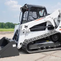 Biała ładowarka gąsienicowa Bobcat T650 z czarną łyżką, widoczna z boku na betonowej powierzchni, w tle drzewa i błękitne niebo.