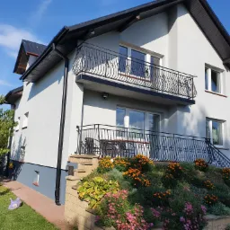 Dwukondygnacyjny dom z jasną elewacją, czarnym dachem i ozdobnymi balkonami, otoczony kwitnącym ogrodem na zboczu