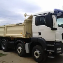 Biała wywrotka MAN TGS 35.440 z beżową skrzynią ładunkową Weiler Kipper, zaparkowana na betonowej nawierzchni przed budynkiem.