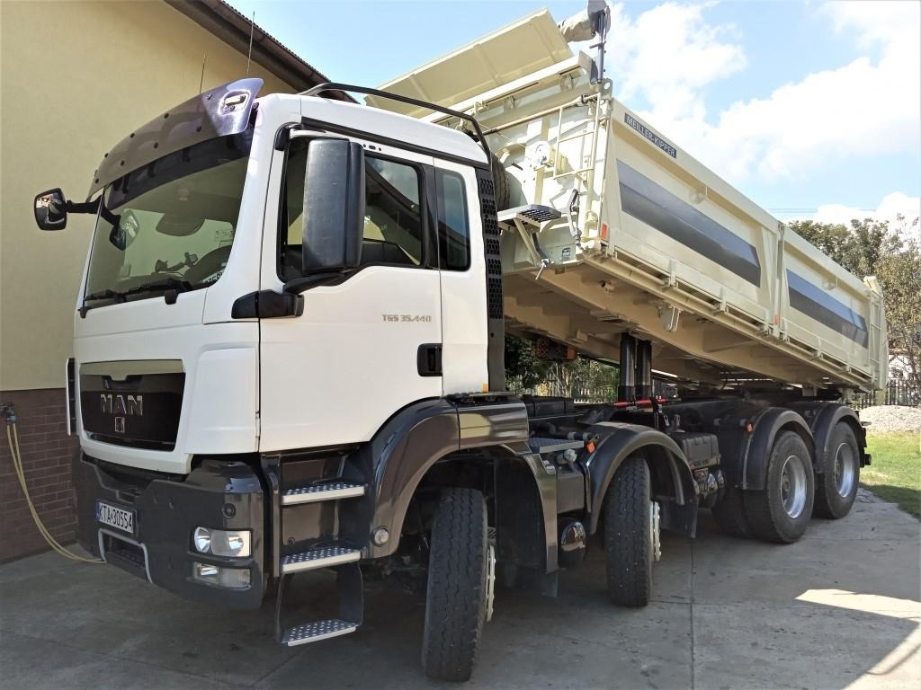 Biała ciężarówka MAN TGS 35.440 z podniesioną skrzynią ładunkową Meiller Kipper, widoczna z przodu i boku na tle budynku i drzew.