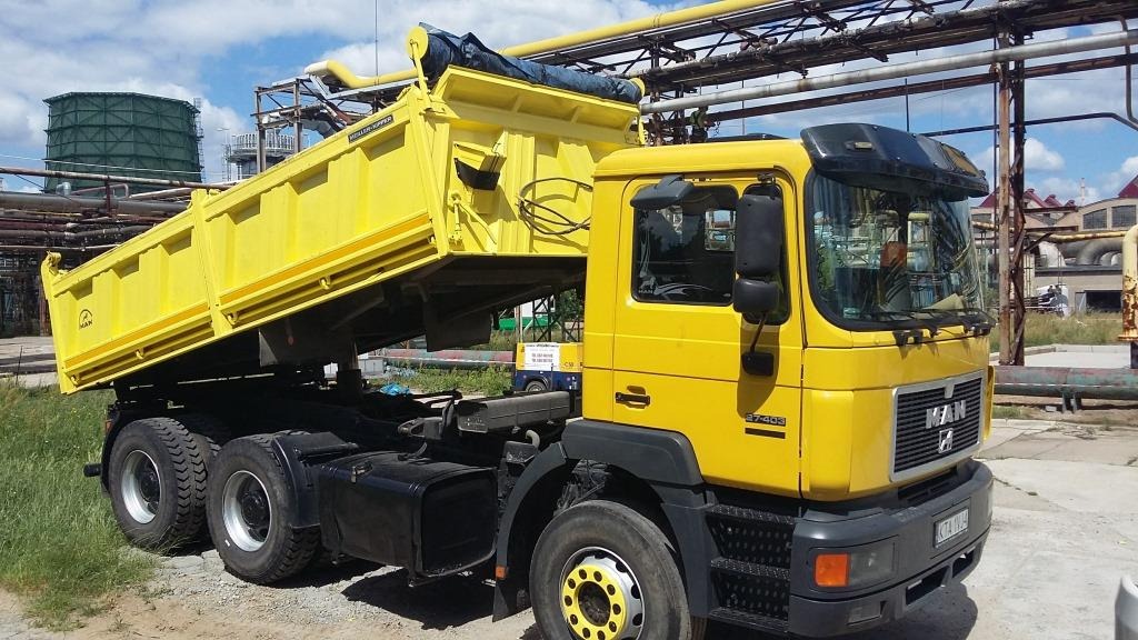 Żółta wywrotka MAN z podniesioną skrzynią ładunkową na tle przemysłowego krajobrazu z rurami i zbiornikami.