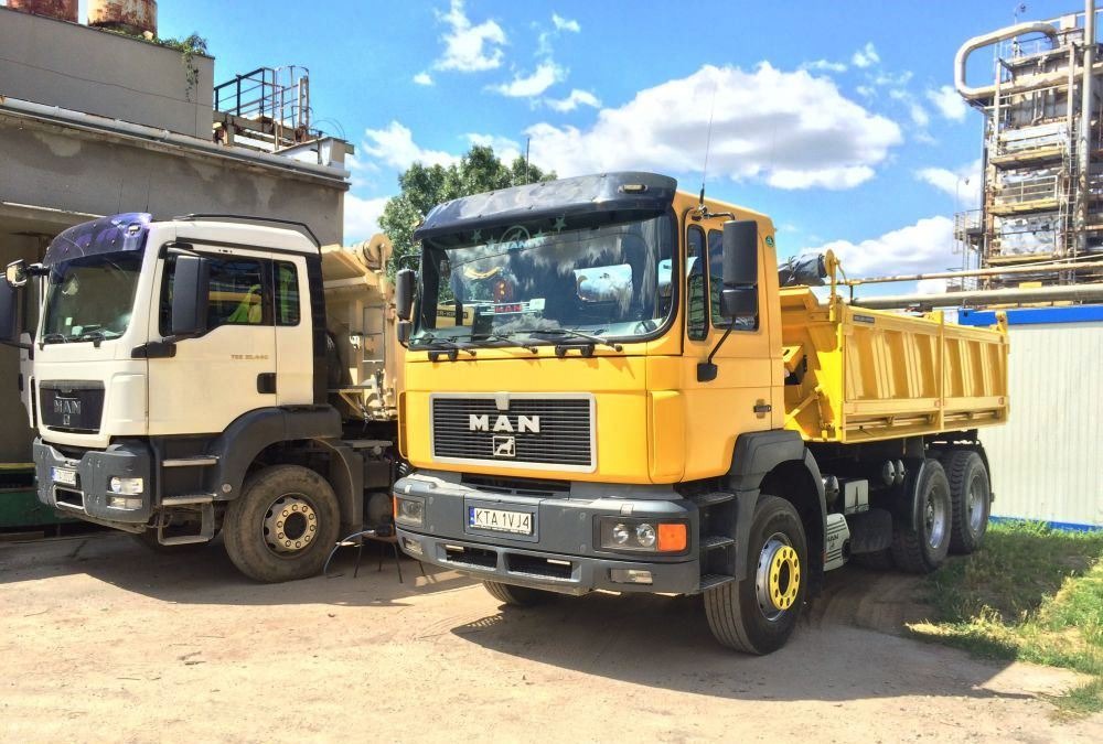 Dwa samochody ciężarowe MAN, żółty i biały, stoją obok siebie na placu budowy, w tle widoczna instalacja przemysłowa i błękitne niebo z chmurami.