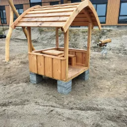 Drewniana altanka z zadaszeniem, ustawiona na betonowych bloczkach, w tle huśtawka typu bocianie gniazdo i drewniany konik na sprężynie, teren przygotowany pod trawnik.