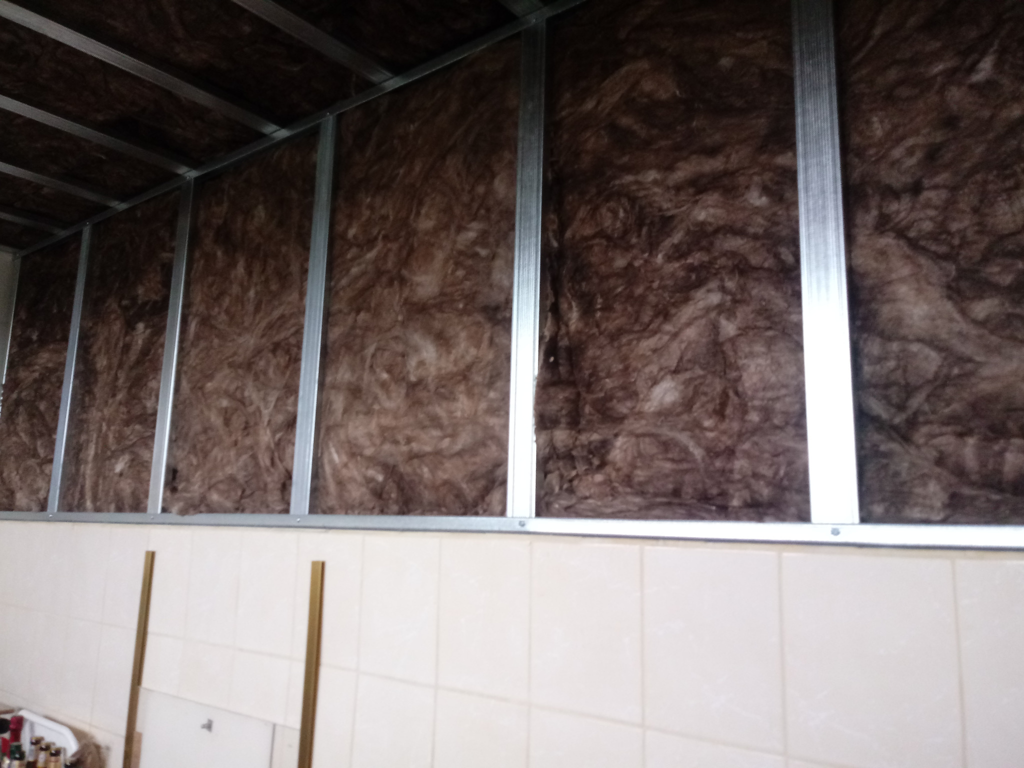 Metalowy stelaż wypełniony brązową wełną mineralną na ścianie wyłożonej jasnymi płytkami ceramicznymi.